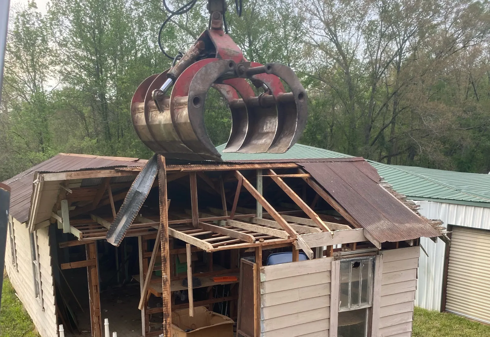 Excavator grapple pulling apart an old frame outbuilding with debris on the ground below, in north Louisiana
