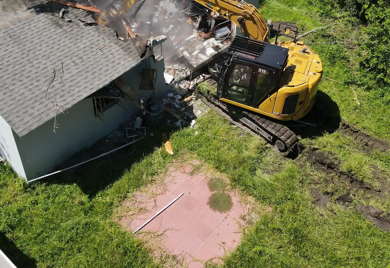 Tracked excavator breaking into the roof and wall of a small house during a teardown, rubble piling up below