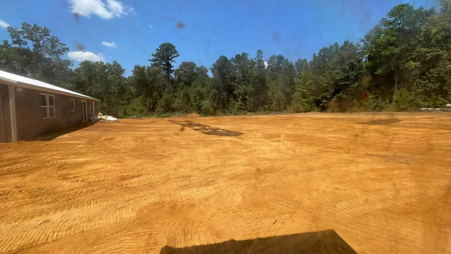 Wide red dirt pad graded out beside a metal building at the edge of the pines