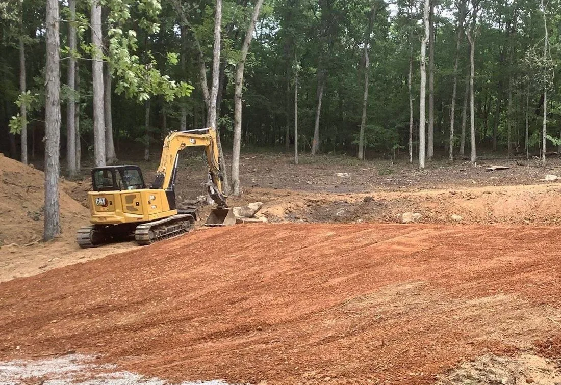 Mini excavator sitting on a freshly cut red dirt pad in a wooded lot