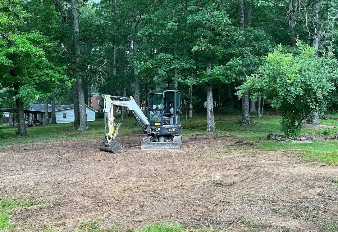Compact excavator on a cleaned-up dirt patch under mature shade trees after an excavation