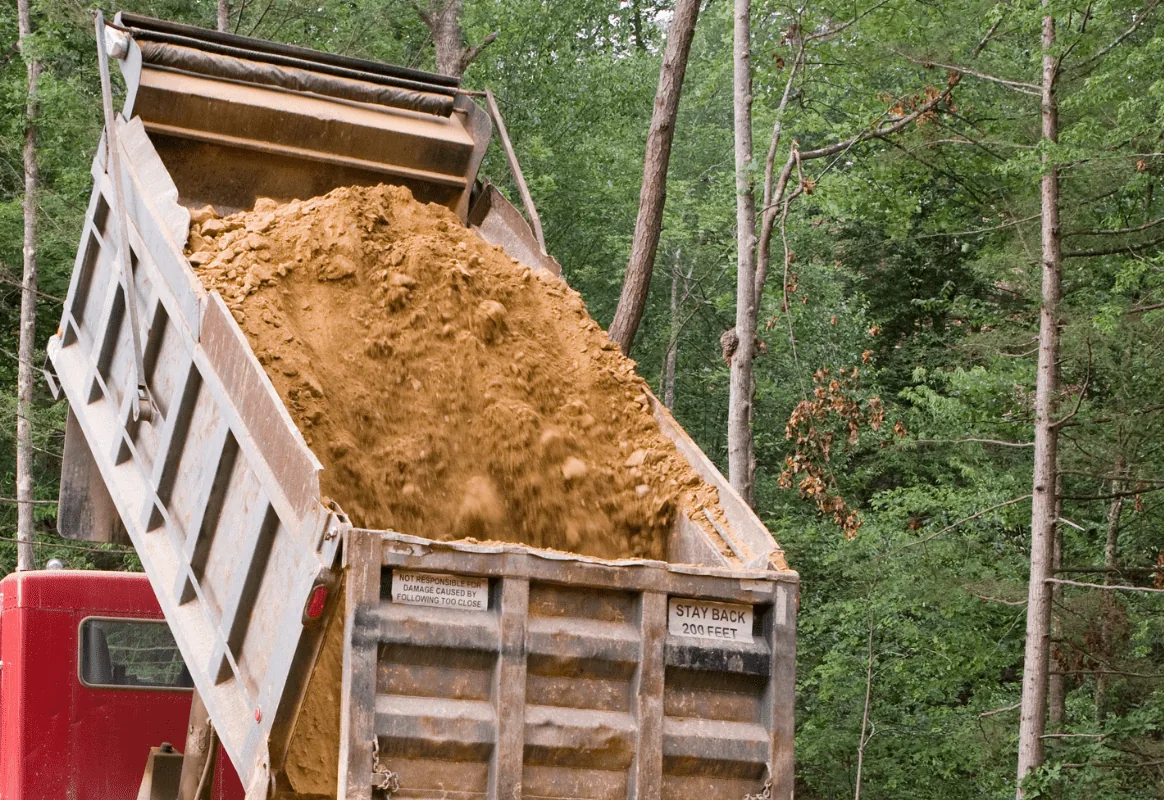 Dump truck bed raised, pouring out a load of tan fill dirt at a wooded job site