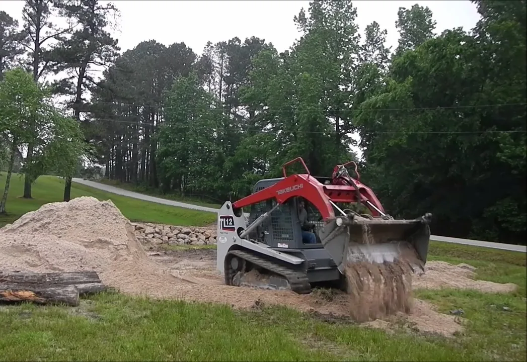 Compact track loader spreading a load of crushed stone along a driveway beside a pine treeline