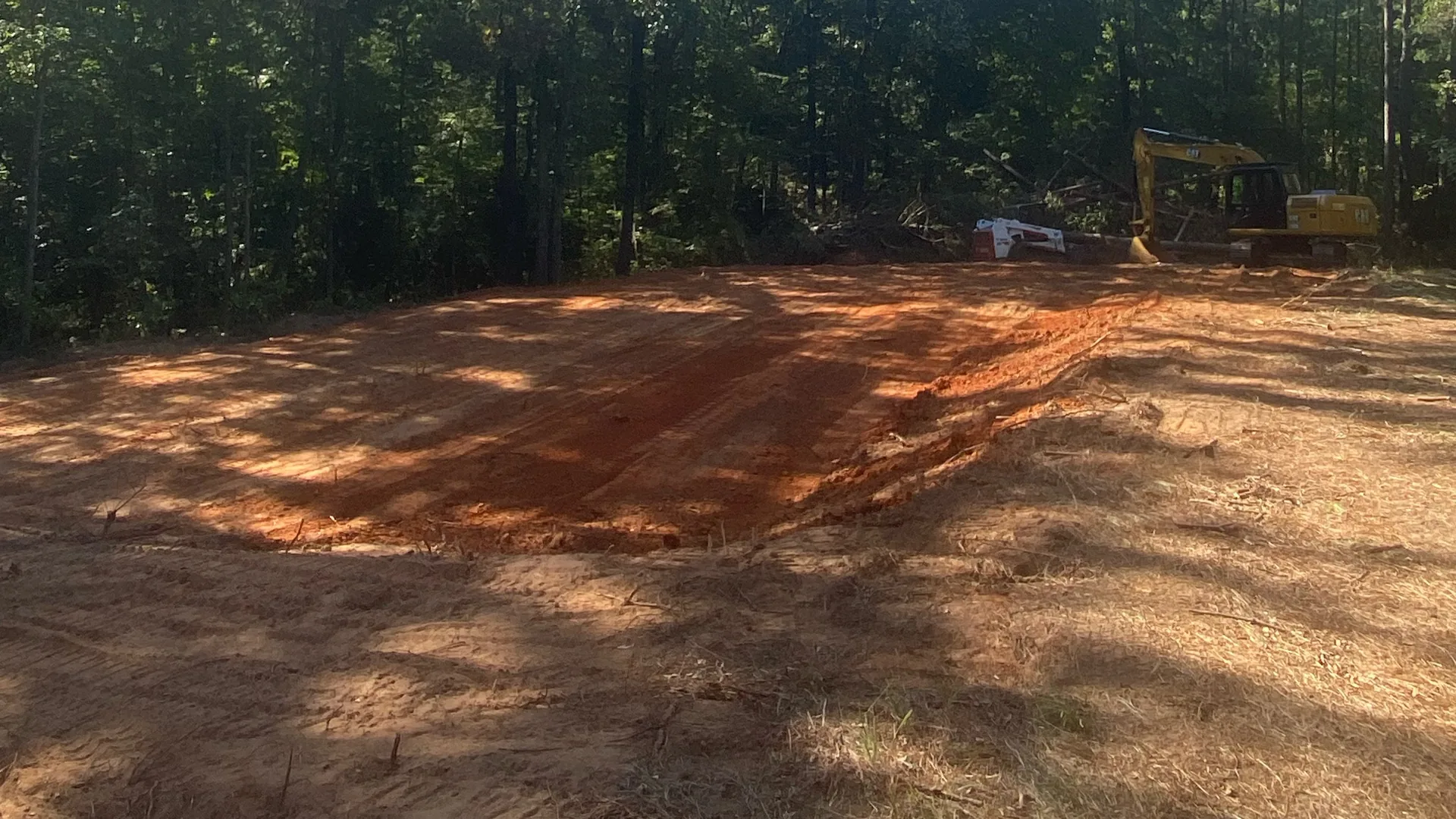 Freshly cleared lot with red dirt spread smooth and a tracked excavator and compact track loader parked at the treeline in north Louisiana