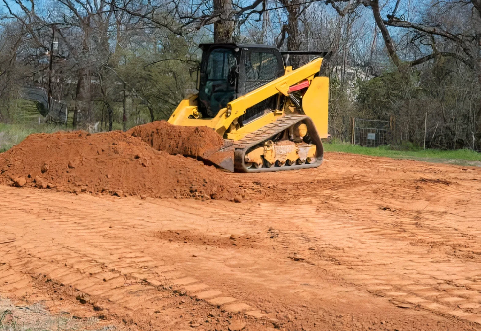 Compact track loader pushing a windrow of red dirt across a lot being graded