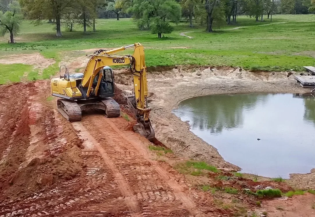 Tracked excavator shaping the red clay dam of a farm pond