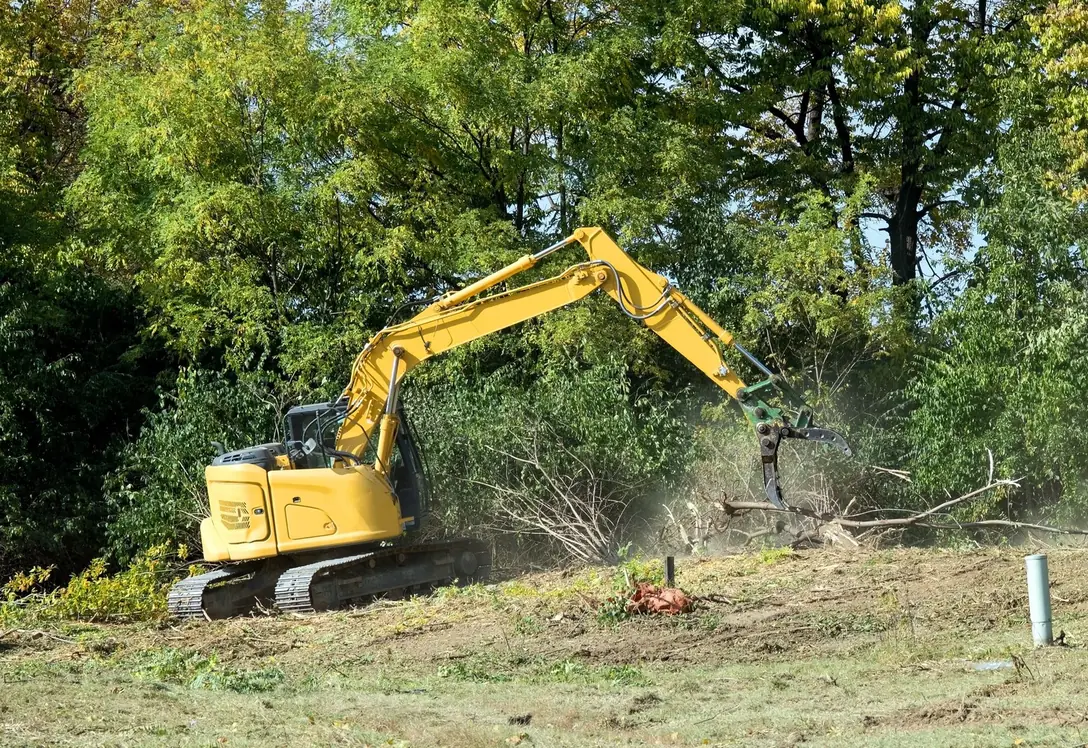 Tracked excavator with a grapple pushing downed brush into a pile at the edge of the woods